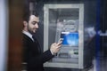 Smiling young businessman standing in front of an ATM and looking at his phone Royalty Free Stock Photo