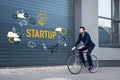 smiling young businessman in suit riding bicycle on street with startup inscription and business icons