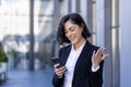 Smiling young businesswoman standing on the street near an office building, using the phone, happy with good news Royalty Free Stock Photo