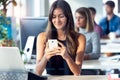 Smiling young businesswoman using her smartphone while working with computer in the modern startup office Royalty Free Stock Photo