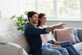 Smiling young caucasian couple sitting on couch, watching tv, husband pointing his finger at screen Royalty Free Stock Photo