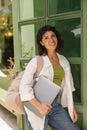 Smiling young caucasian woman looking at camera, holding backpack, laptop outdoors.