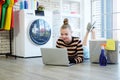 Smiling young Caucasian woman relaxing with laptop for online social media while waiting for the washing machine to finish