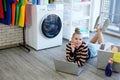 Smiling young Caucasian woman relaxing with laptop for online social media while waiting for the washing machine to finish