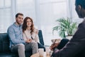 Smiling young couple sitting on sofa and talking to counselor with pencil Royalty Free Stock Photo