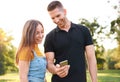 Smiling young couple using smartphone outdoor in sunset - Happy friends having fun and checking social media together in a park - Royalty Free Stock Photo