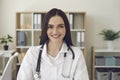 Smiling young doctor looking at camera ready for online consultation in her modern hospital office