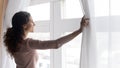 Smiling young dreamy woman opening curtains in morning.