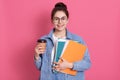 Smiling young European student girl holds folders wears denim jacket. Pleased female with dark hair and hair bun drinks coffee,