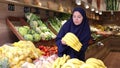 Smiling young european woman in traditional islamic dress and khimar choosing ripe sweet bananas while shopping at fruit