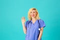 Smiling young female doctor or nurse wearing blue scrubs and stethoscope, waving her hand at camera Royalty Free Stock Photo