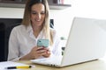 Smiling young female entrepreneur reading emails on her cellphone while working on a laptop at home. Telecommuting lifestyle Royalty Free Stock Photo