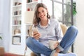 Smiling young female sitting in the armchair in the living room and using with her mobile phone Royalty Free Stock Photo