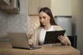 A smiling young freelance woman is sitting at a table in her kitchen, taking notes and working on a laptop Royalty Free Stock Photo