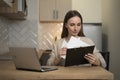 A smiling young freelance woman is sitting at a table in her kitchen, taking notes and working on a laptop Royalty Free Stock Photo