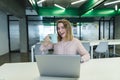 Smiling young girl with a cup of coffee in her arms sitting at the desk in the office and using a laptop Royalty Free Stock Photo