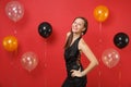 Smiling young girl in little black dress celebrating standing with arms akimbo on bright red background air balloons. St Royalty Free Stock Photo