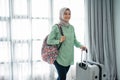 Smiling young hijab traveler carrying her bag and holding suitcase