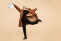 Smiling young hipster woman with blonde short hair wearing a coat and sunglasses dance with clock isolated over beige Royalty Free Stock Photo