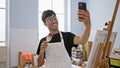 Smiling young hispanic man artist taking a splendid selfie picture with his smartphone at the bustling art studio, canvas in one Royalty Free Stock Photo