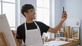 Smiling young hispanic man artist taking a splendid selfie picture with his smartphone at the bustling art studio, canvas in one Royalty Free Stock Photo