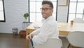 Smiling young hispanic man joyfully sits at office table, exuding confidence with business success as a hardworking worker