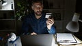 Smiling young hispanic man, laptop at hand and a credit card at work, late-night shopping spree at the office Royalty Free Stock Photo