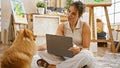 Smiling young hispanic woman artist, with her dog, sitting on studio floor, engrossed in painting lesson on laptop Royalty Free Stock Photo