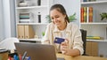 Smiling young hispanic woman, a beautiful business worker, enjoying success while shopping online with credit card on laptop at Royalty Free Stock Photo