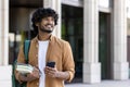 Smiling young Indian male student standing outside campus, holding books, backpack and using phone. Looks to the side Royalty Free Stock Photo