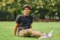 Smiling young indian man candid portrait in black t shirt, silver neck chain sitting on green lawn Royalty Free Stock Photo