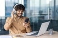 Smiling young Indian man sitting at desk in office with laptop and tablet, wearing white headphones, using mobile phone Royalty Free Stock Photo