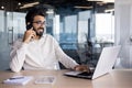 A smiling young Indian man is sitting at a desk in the office, using a laptop and talking on the phone Royalty Free Stock Photo