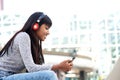 Smiling young Indian woman sitting outside in the city listening to music with mobile phone and headphones Royalty Free Stock Photo