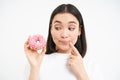 Smiling young korean woman, thinking and gazing at pink glazed doughnut, white background