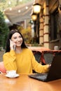 smiling young lady in orange sweater. businesswoman talking on mobile phone while working on laptop remotely in autumn Royalty Free Stock Photo