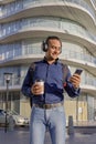 Smiling young latin man with headphones looking at his mobile phone and holding a paper cup with coffee in his hand Royalty Free Stock Photo