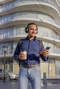 Smiling young latin man with headphones looking at his mobile phone and holding a paper cup with coffee in his hand in a city Royalty Free Stock Photo