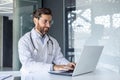 A smiling young male doctor is working in the office of the clinic using a laptop. Sitting in a white coat at the desk Royalty Free Stock Photo