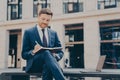 Smiling young man dressed in blue suit taking notes in notebook while spending time outdoors Royalty Free Stock Photo