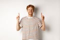 Smiling young male student with red hair showing logo, pointing fingers up and looking happy, standing over white Royalty Free Stock Photo