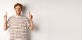 Smiling young male student with red hair showing logo, pointing fingers up and looking happy, standing over white Royalty Free Stock Photo