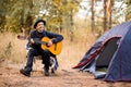 Smiling young man in black hat sitting near touristic tent and playing guitar in forest Royalty Free Stock Photo
