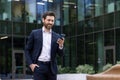 A smiling young man in a business suit is walking near an office building rubbing his hand in his pocket and using a Royalty Free Stock Photo