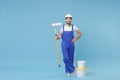 Smiling young man in coveralls protective helmet hardhat hold paint roller, bucket isolated on pastel blue wall Royalty Free Stock Photo