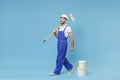 Smiling young man in coveralls protective helmet hardhat hold paint roller, bucket isolated on pastel blue wall Royalty Free Stock Photo