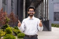 Confident young businessman with glasses giving thumbs up outside modern office building, expressing approval Royalty Free Stock Photo