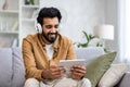 Smiling young man in headphones sits on the sofa in the living room, holds a tablet in his hands, listens to his Royalty Free Stock Photo