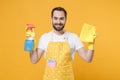 Smiling young man househusband in apron rubber gloves hold spray with washing cleanser, cleaning rag while doing