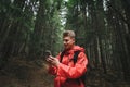 Smiling young man in a red raincoat stands on a forest path in the mountains and uses a smartphone with a positive face. Happy